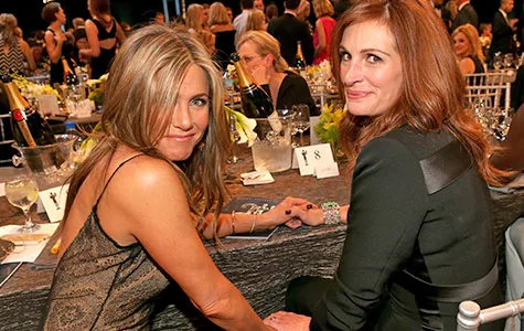 Two women seated at a formal event table, smiling back at the camera, surrounded by guests and champagne bottles.