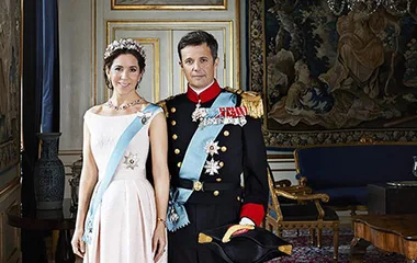Royals in formal attire: a woman in a light gown and tiara stands beside a man in a military uniform with sash and hat.