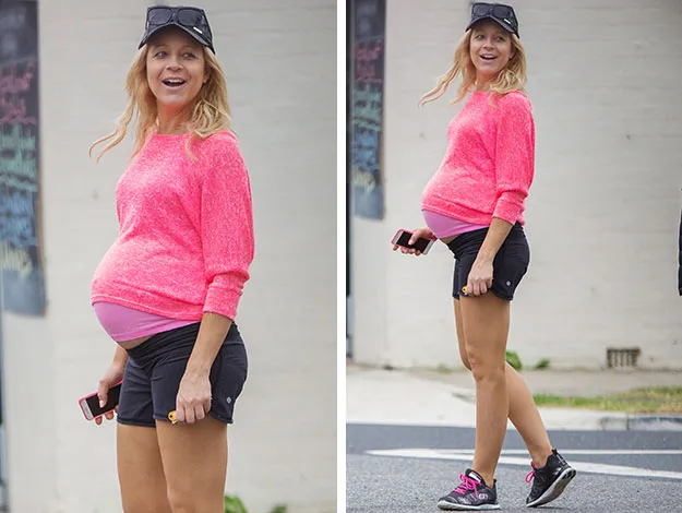 Pregnant woman in pink sweater, black shorts, and cap walking outdoors, smiling, holding a phone.