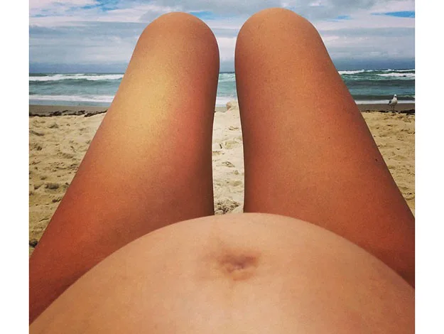 View of a pregnant belly and legs at the beach with ocean in the background.