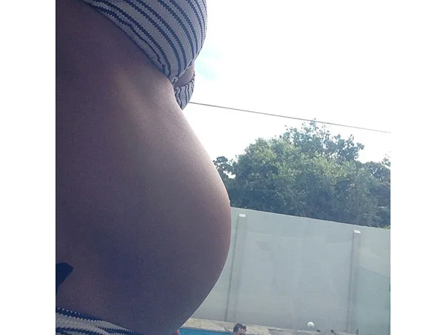 Pregnant woman in striped bikini stands by a pool with trees in the background.