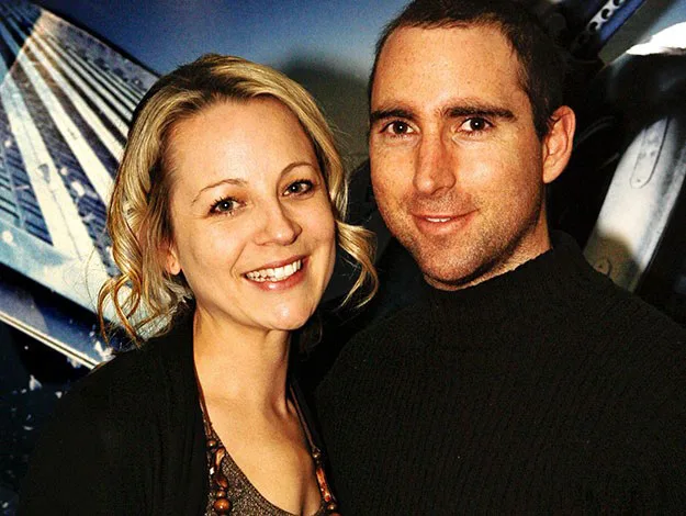 A smiling woman with blonde hair and a man in a black sweater pose together in front of a futuristic building background.