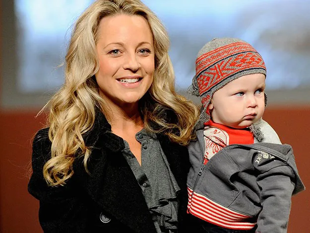 Woman with long blonde hair holding a baby wearing a red and gray knit hat.