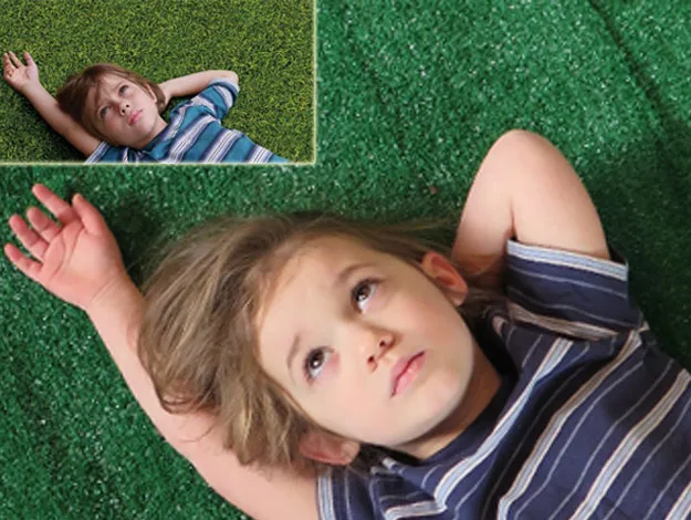 A child with long hair lies on green grass, looking contemplative, wearing a blue striped shirt.