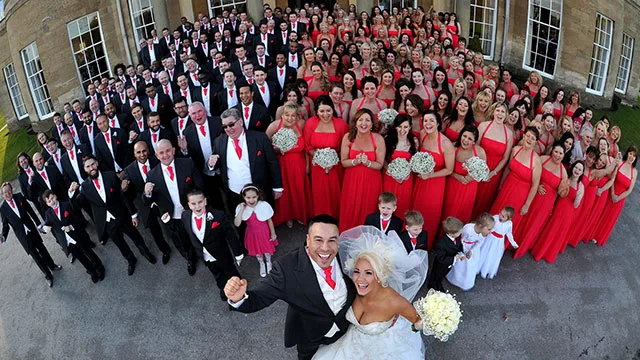 Bride and groom with 130 bridesmaids and guests in matching red and black outfits posing outside a large building.