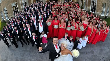 Bride and groom with 130 bridesmaids and guests in matching red and black outfits posing outside a large building.
