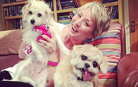 A person smiling and holding two small dogs, one wearing a pink bandana, sitting on a sofa with bookshelves in the background.