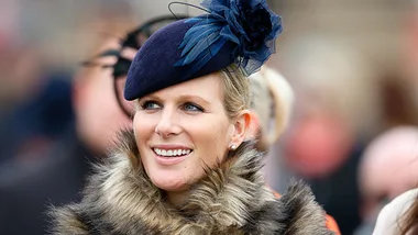 A smiling woman in a fur coat and blue hat with a flower detail, standing outdoors.