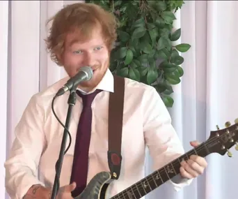 Musician with guitar and mic, performing in front of white curtain and greenery.