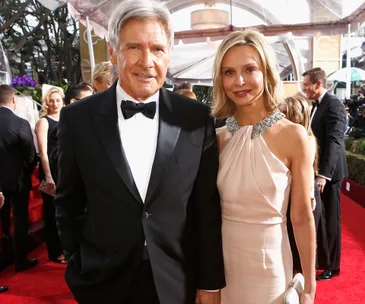 Man in a tuxedo and woman in a white gown on a red carpet, surrounded by other guests.
