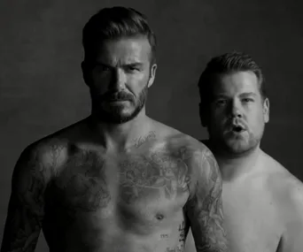 Two men standing shirtless; one is tattooed and serious, the other is looking amused in the background.