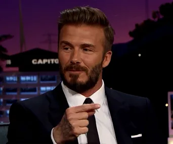 David Beckham in a suit on a talk show, gesturing with his hand.