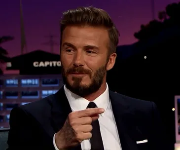 David Beckham in a suit on a talk show, gesturing with his hand.