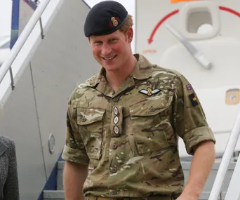 Prince Harry in military uniform, smiling while descending airplane stairs.