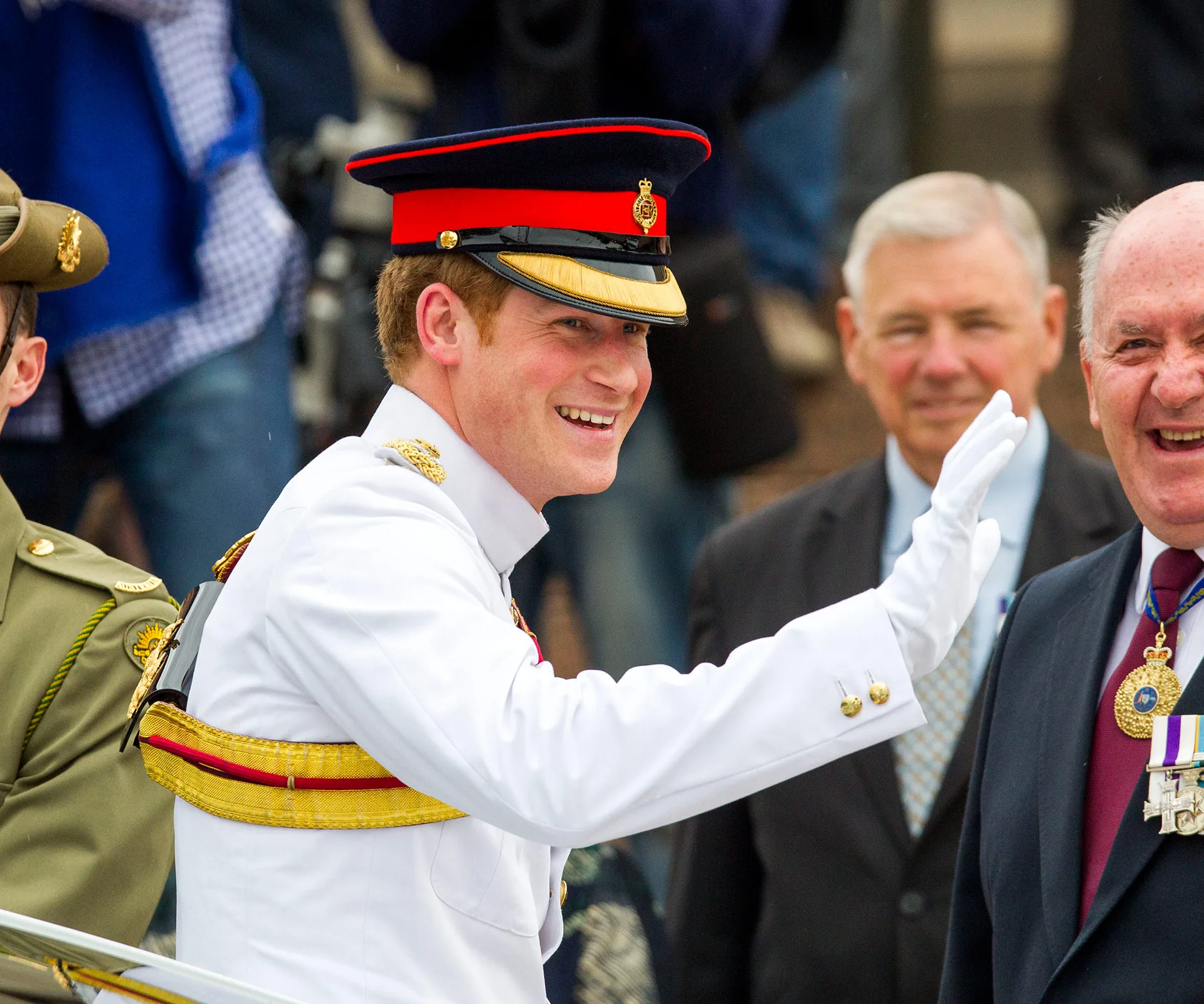 Prince Harry in uniform, smiling and waving at the National War Memorial in Canberra, with people in the background.