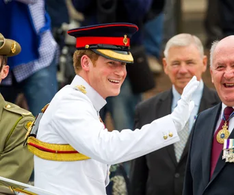 Prince Harry in uniform, smiling and waving at the National War Memorial in Canberra, with people in the background.