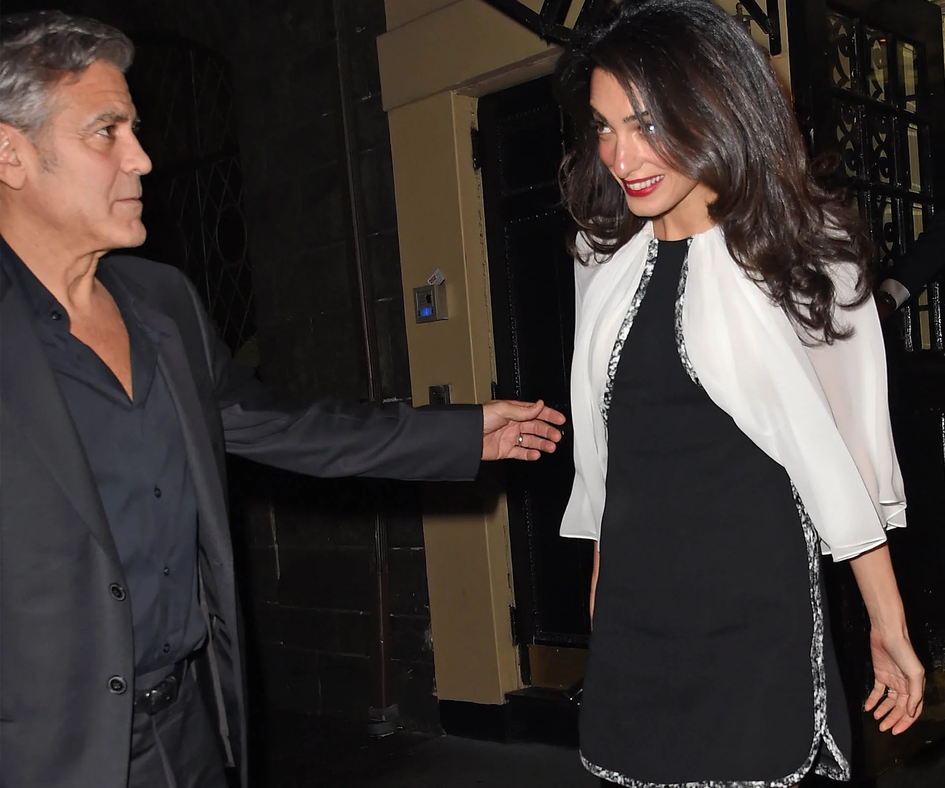 Man in suit and woman in black dress with white shawl smiling outside a building at night.