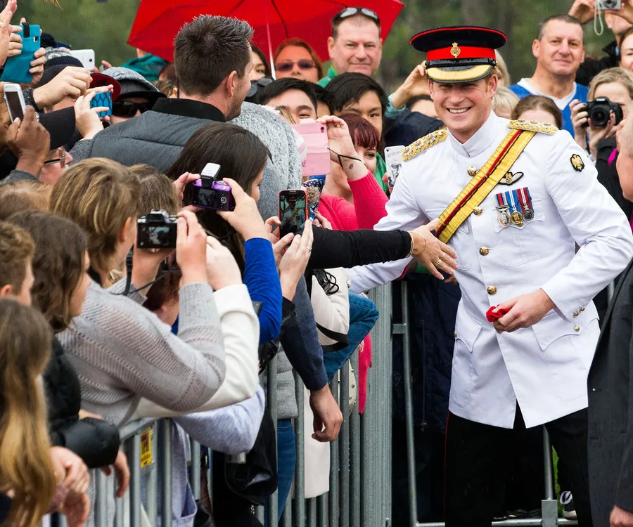 Prince in military uniform smiles and greets a crowd holding cameras and phones for selfies.