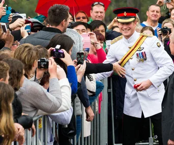 Prince in military uniform smiles and greets a crowd holding cameras and phones for selfies.