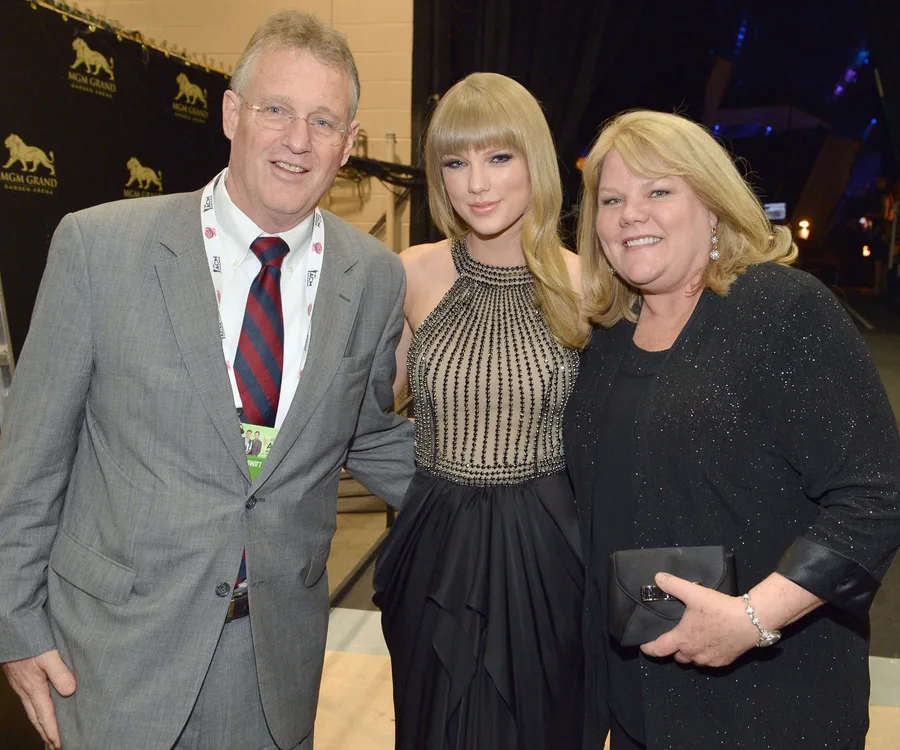 A woman in a shimmering dress poses with a smiling man in a suit and a woman in black, against a backdrop.