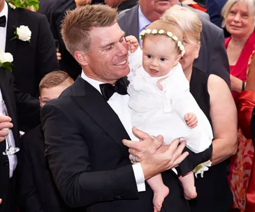 "Man in a tuxedo holds a smiling baby wearing a white dress and flower headband at a formal event."
