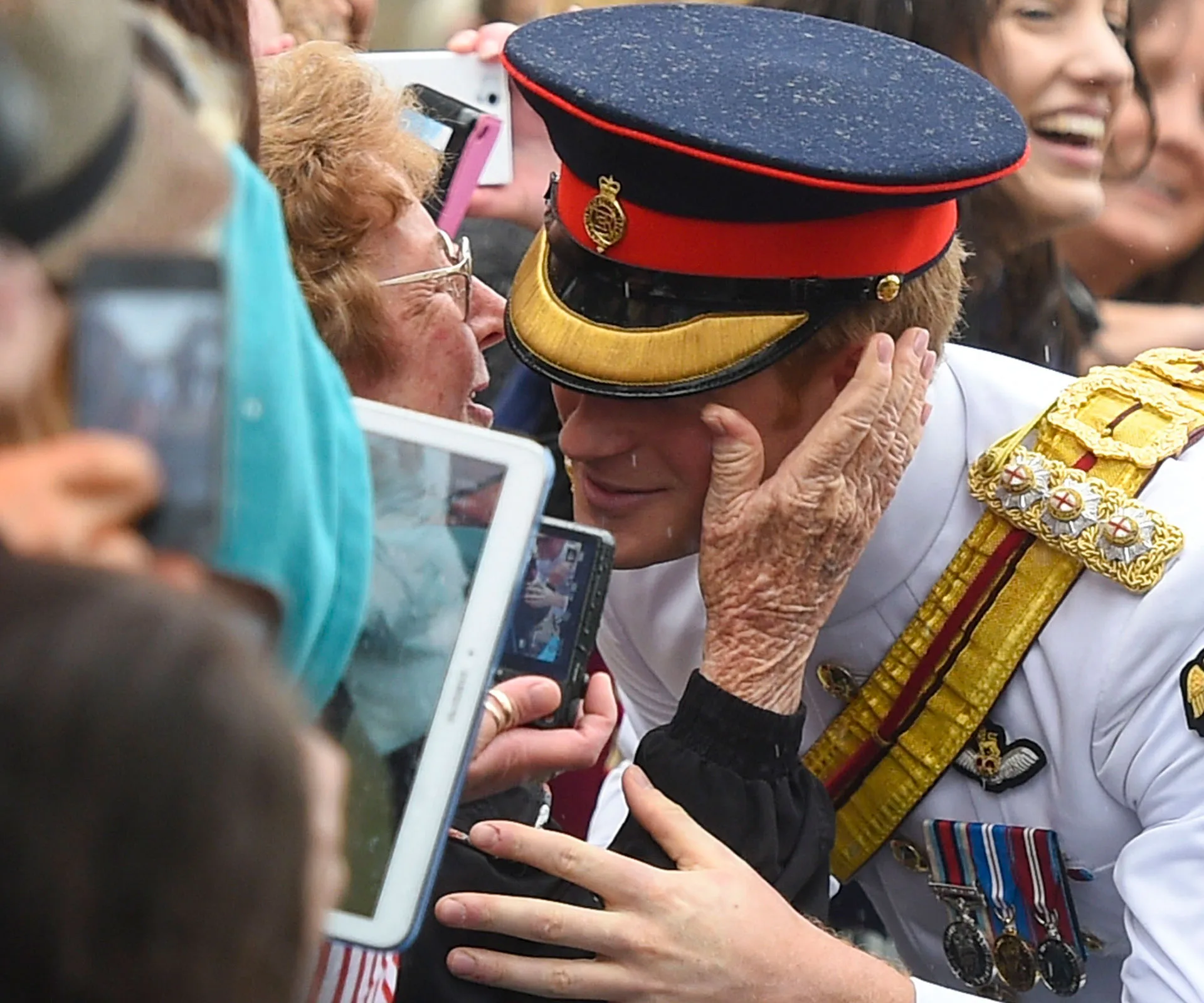 Kissing Prince in uniform, elderly woman holding his face, surrounded by onlookers with phones.