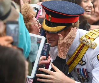Kissing Prince in uniform, elderly woman holding his face, surrounded by onlookers with phones.