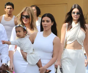 Mom carrying toddler in white dress with flower headband; surrounded by women in white outfits.