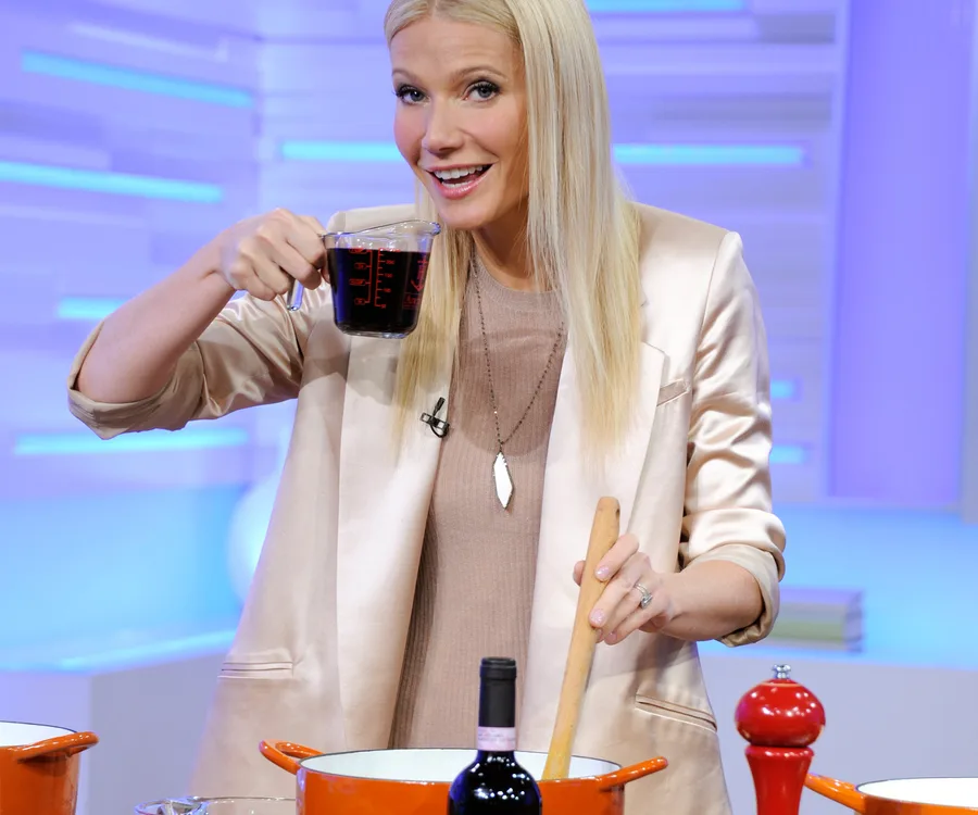 A woman smiling, holding a measuring cup, and stirring a pot in a kitchen setting.