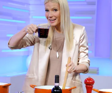 A woman smiling, holding a measuring cup, and stirring a pot in a kitchen setting.