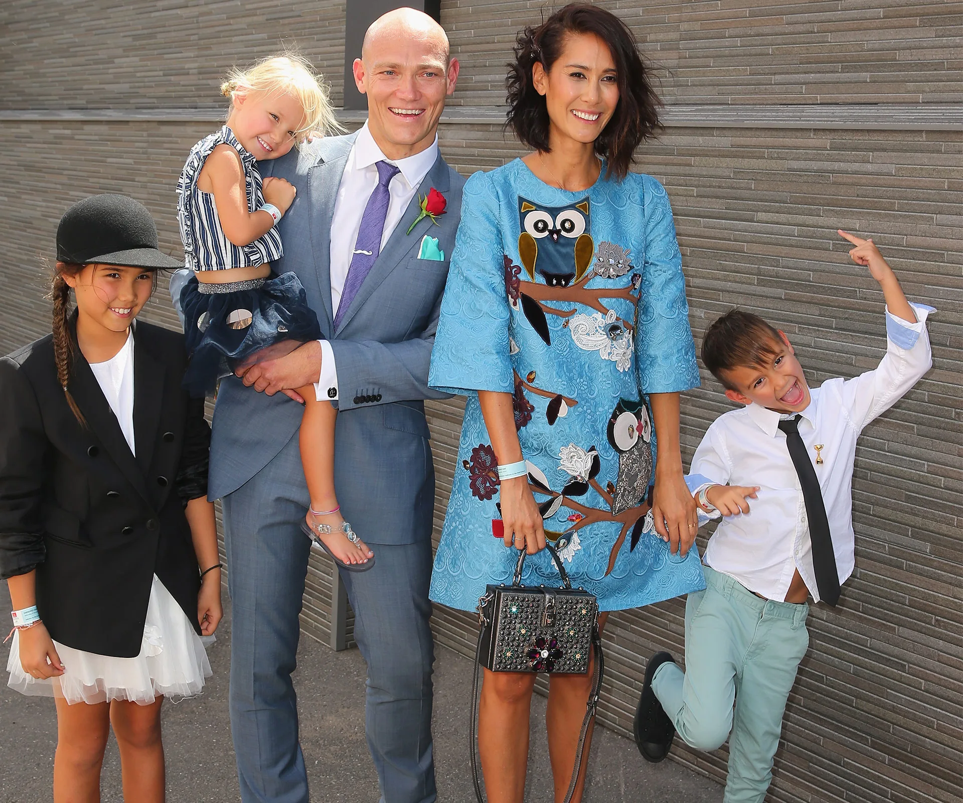 Family posing together, man in blue suit holding child. Woman in blue dress, two other children, all smiling.