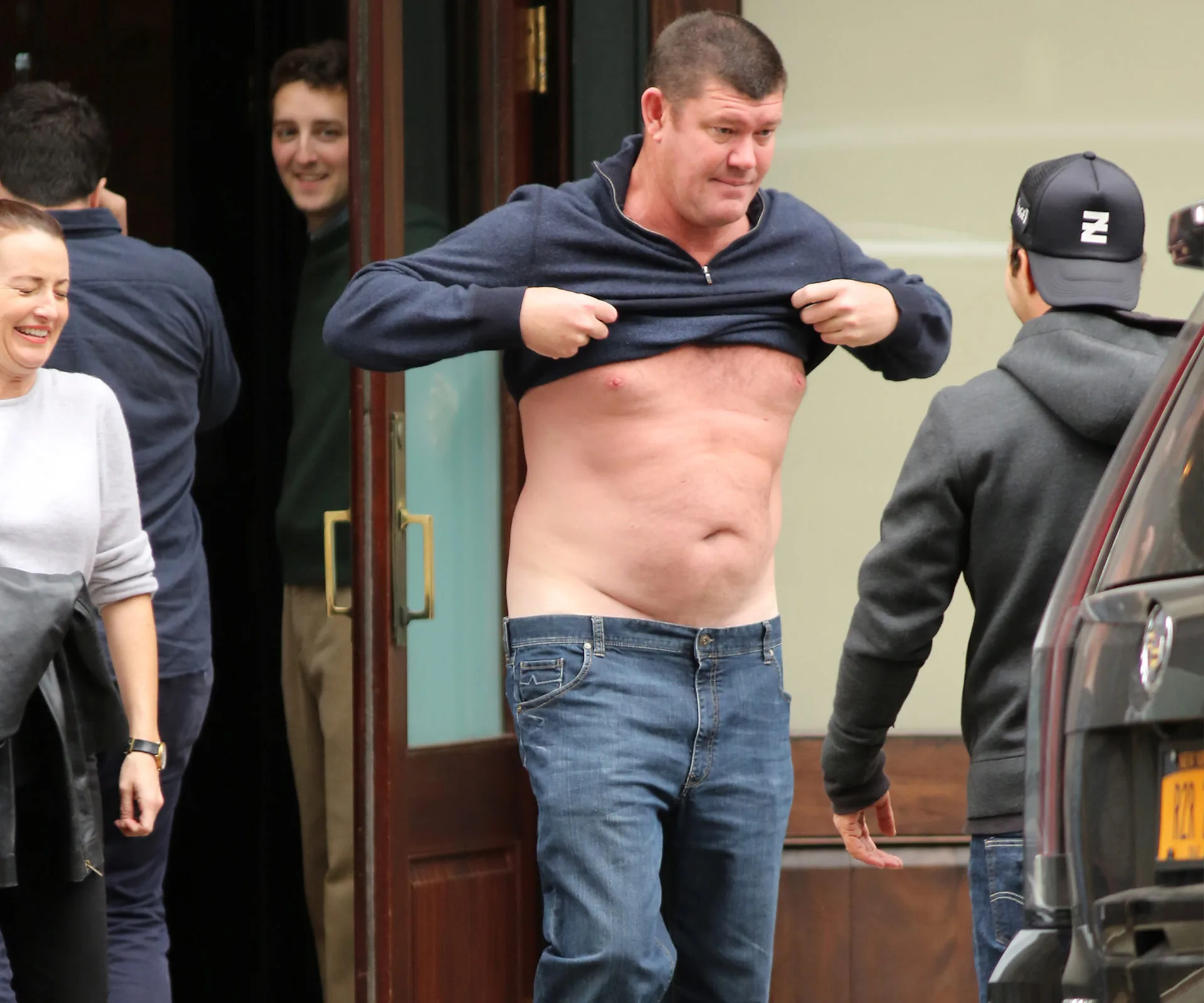 Man lifting his shirt outdoors, with several people around him laughing and looking on.