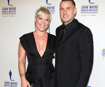 A smiling couple at an event, woman in a black dress, man in a suit, backdrop has "John Wayne Cancer Institute" logo.