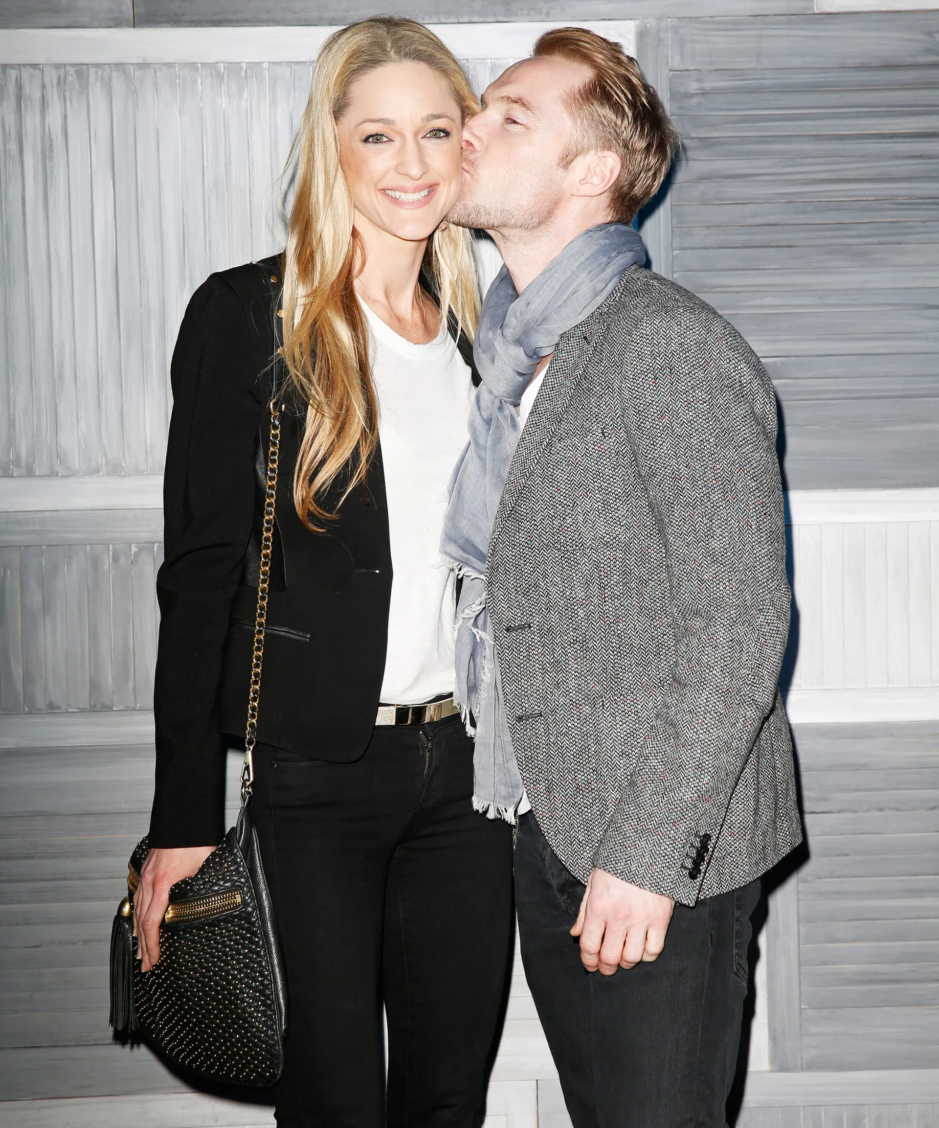 A couple poses, smiling with affection; man kisses woman on cheek, embracing against a textured background.