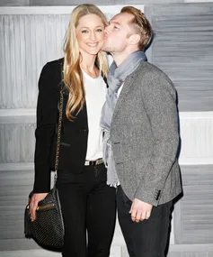 A couple poses, smiling with affection; man kisses woman on cheek, embracing against a textured background.