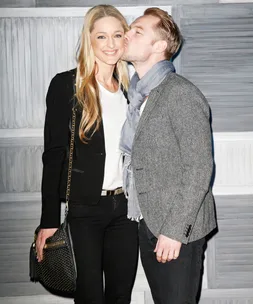 A couple poses, smiling with affection; man kisses woman on cheek, embracing against a textured background.