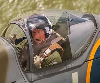 Pilot in cockpit giving a thumbs-up from inside a fighter jet, wearing a helmet and headset.
