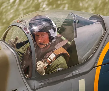 Pilot in cockpit giving a thumbs-up from inside a fighter jet, wearing a helmet and headset.