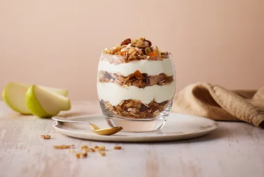 A yogurt parfait with granola and fruit layered in a glass, next to sliced pear on a plate with a spoon.