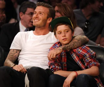 A man in a white shirt smiles while sitting with a child in a plaid shirt and green cap in a crowded stadium.