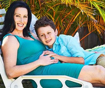 Woman in a blue dress reclines on a lounge chair outdoors, with a young boy resting his head on her belly.