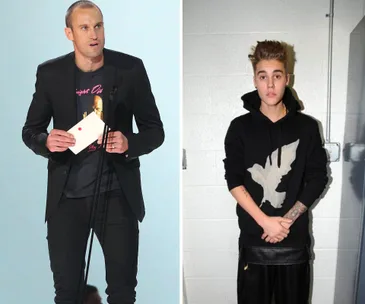 Two individuals, one speaking at a podium holding a card, the other in a black hoodie with a bird design standing against a wall.