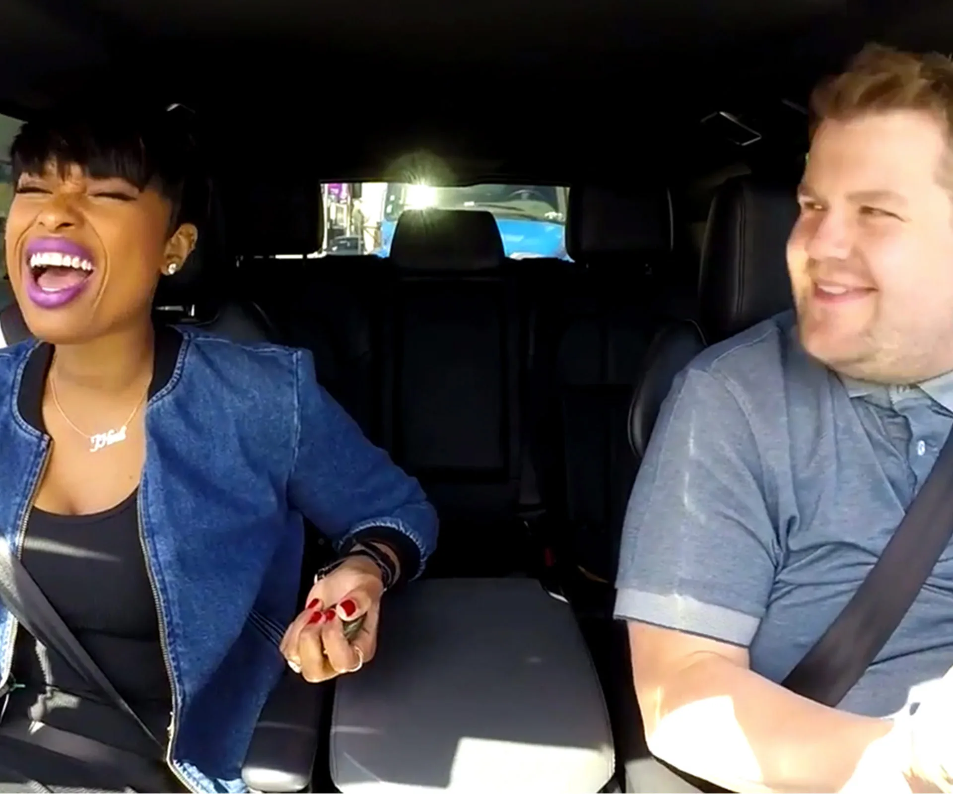 Car interior with two people smiling, one wearing a denim jacket, the other a gray shirt, both seatbelted.