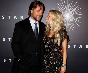 A man in a suit and a woman in a floral dress smiling at each other on a red carpet event.