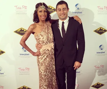 A woman in an elegant dress and a man in a suit pose at a fundraiser event, with sponsor logos in the background.