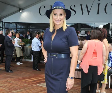 A woman in a blue dress and hat smiles at a social event with people mingling in the background.