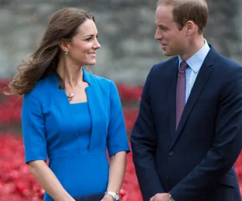 A couple stands outdoors, smiling at each other; the woman wears a blue dress, and the man is in a suit.