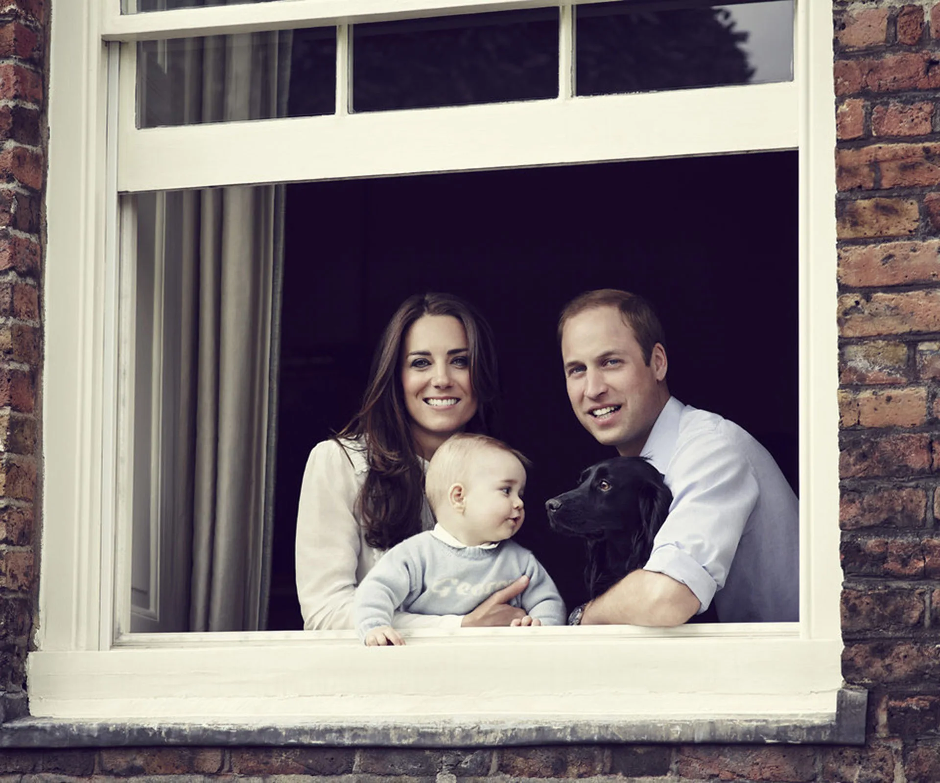 A family of three with a baby and a dog gazing out from a window, smiling.