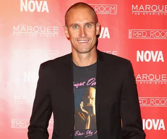 Headshot of a smiling man in a black blazer, standing against a red backdrop with logos, at an event.