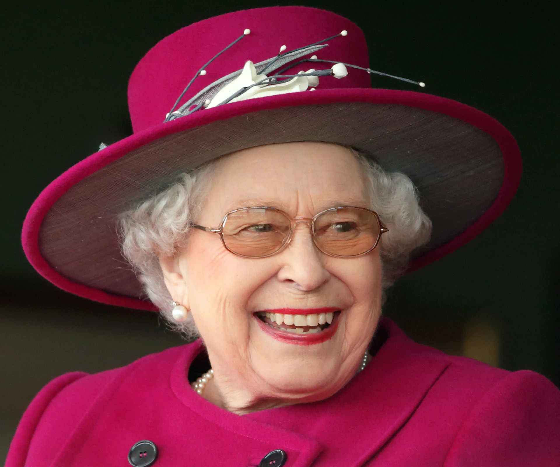 Smiling older woman in a bright magenta coat and hat with decorative elements.